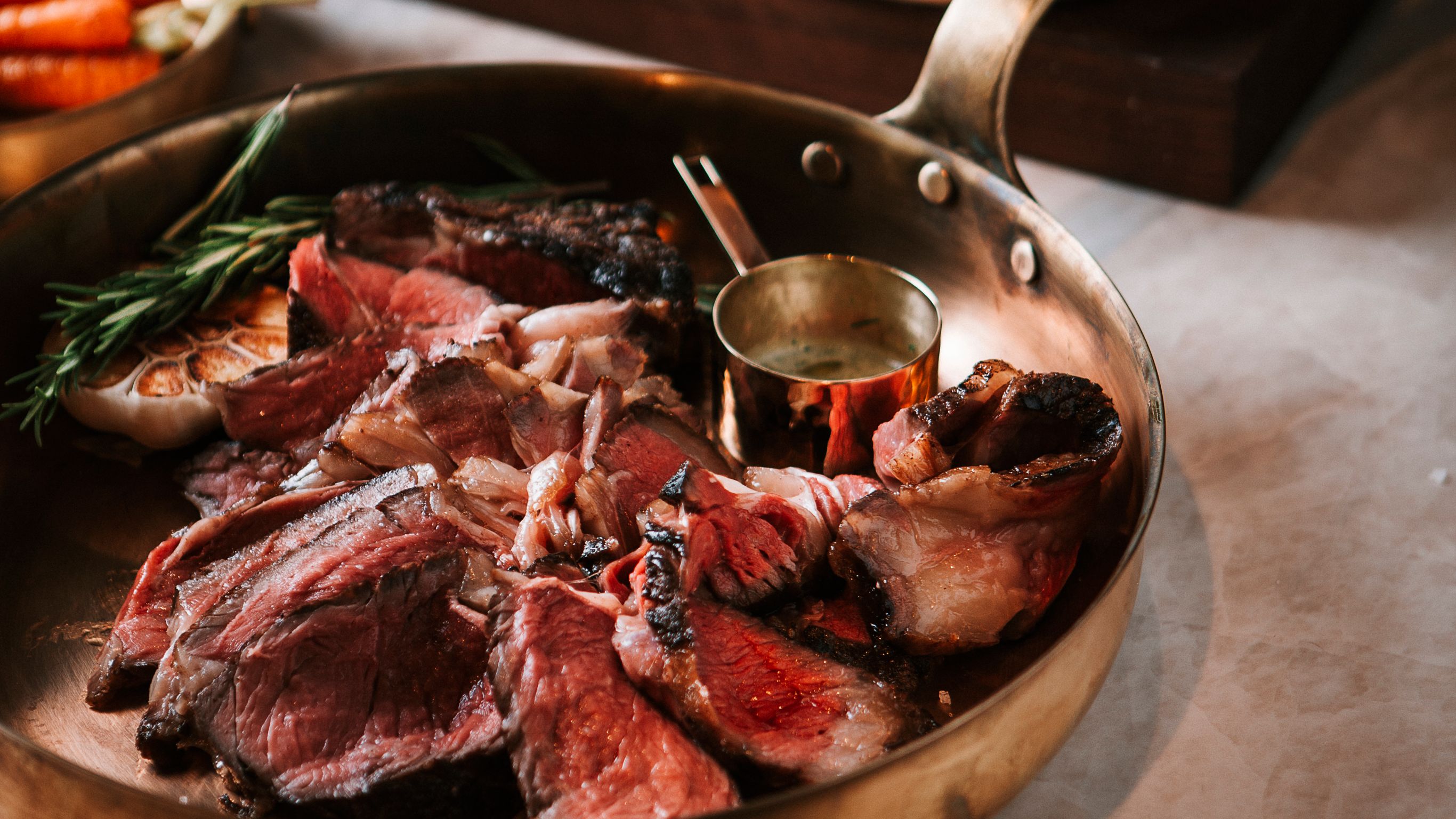 Pan with sliced meat, herbs, and garlic, accompanied by a small container of sauce.